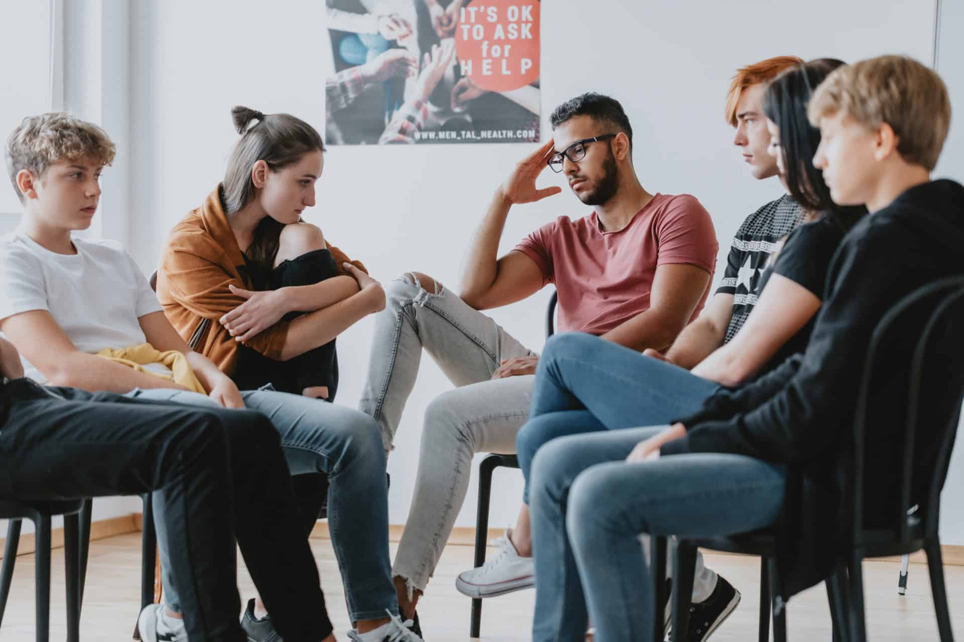 A group of six people sit in a circle during a discussion session in a room. A poster on the wall reads "It's OK to ask for help.