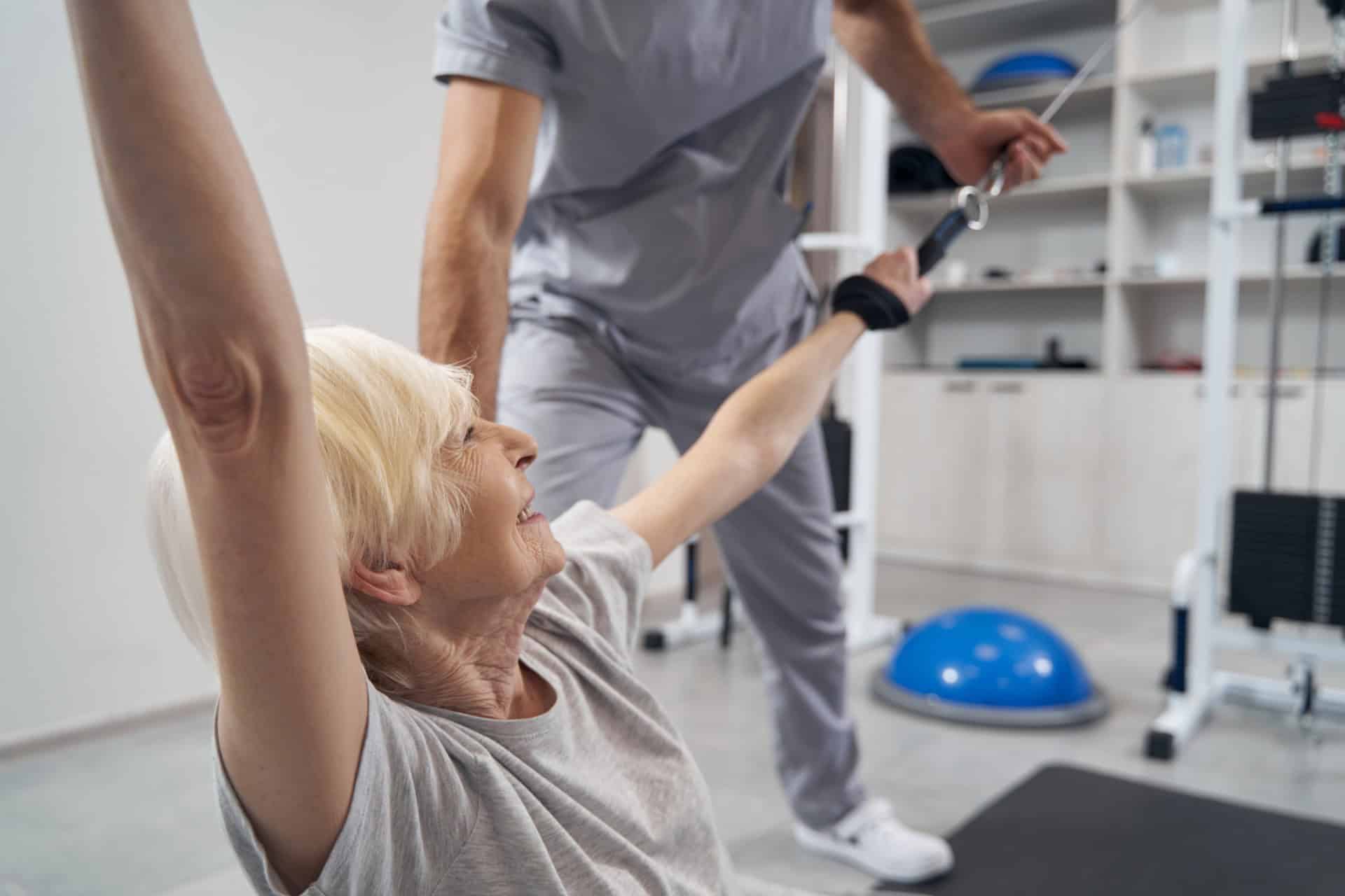 An elderly person in a gym is assisted by a trainer while exercising with resistance bands.