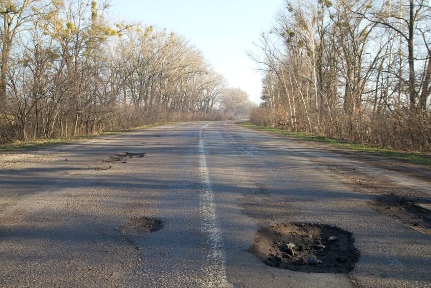 A rural road with visible potholes runs through leafless trees under a clear sky.