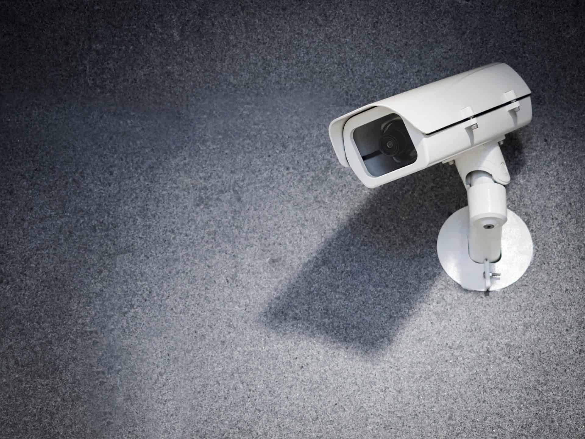 A white security camera mounted on a gray wall with its shadow visible.
