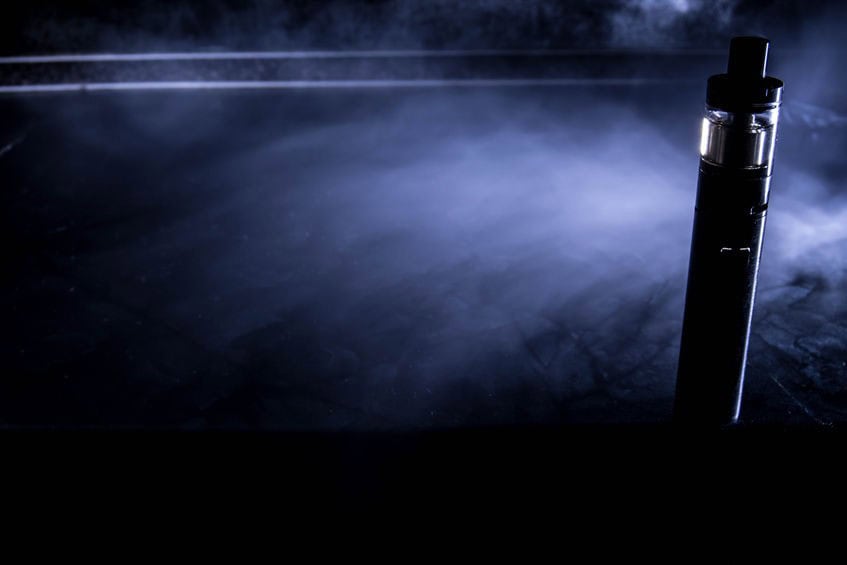 A vape pen stands upright on a dark surface surrounded by smoke in a dimly lit environment.