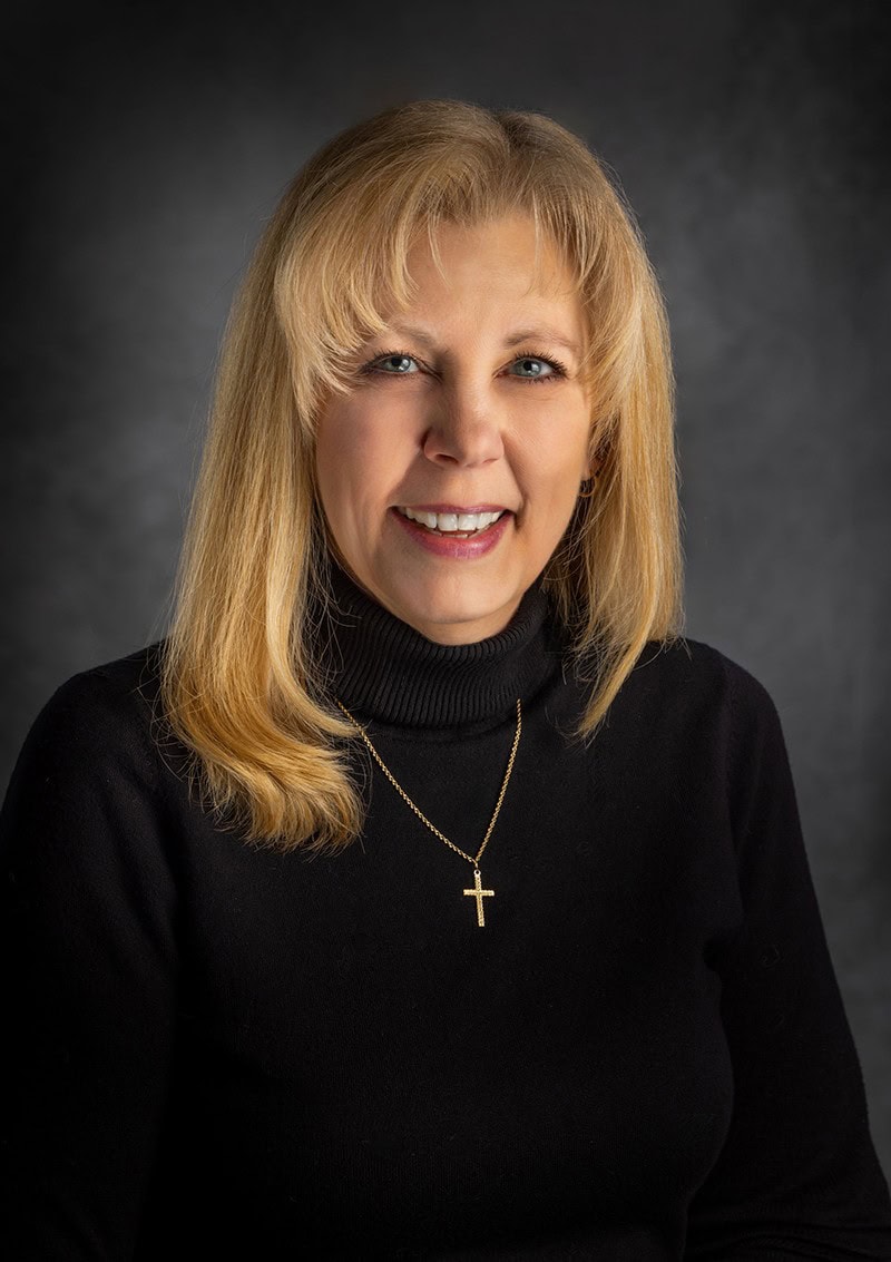 A woman with long blonde hair wearing a black turtleneck and a cross necklace smiles against a dark background.