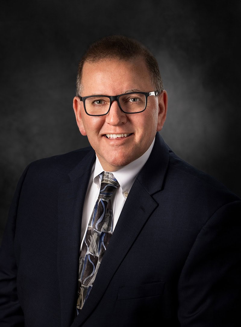 Smiling man in a suit and tie with glasses, set against a dark, plain background.