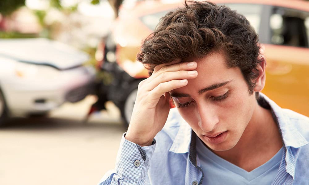 A person looks distressed, holding their forehead, with two cars involved in a rear-end collision in the background.