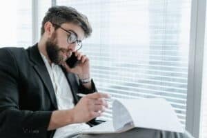 Man with glasses sitting by a window, talking on a phone, and looking at documents.
