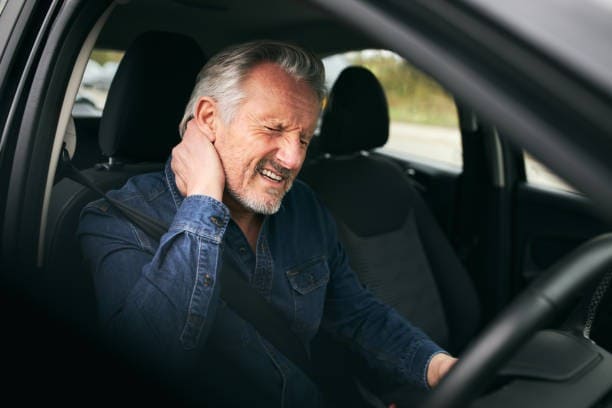 Older man in a car driver's seat rubbing his neck with a pained expression, wearing a denim shirt.