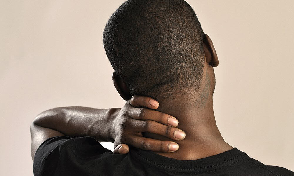 Person holding the back of their neck with one hand, as if in discomfort or pain. Background is plain.