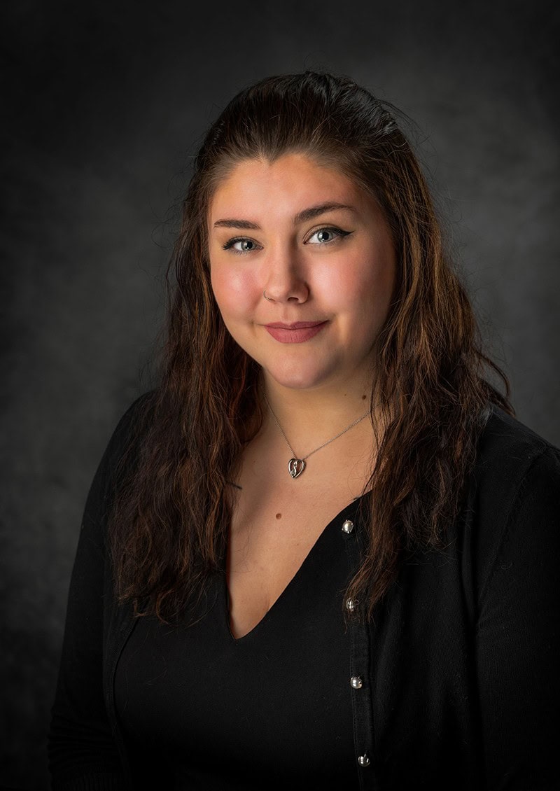 Person with long brown hair wearing a black top, pictured against a dark background.