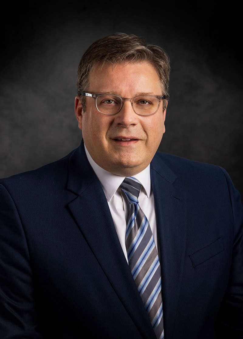 Man in a suit and striped tie against a dark background, wearing glasses and looking at the camera.