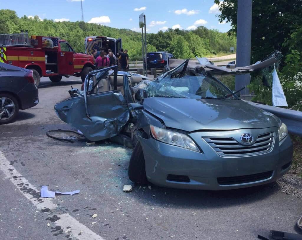 Damaged gray car with crushed roof and shattered windshield on roadside. Emergency responders and fire truck in background. Clear sky and trees visible.
