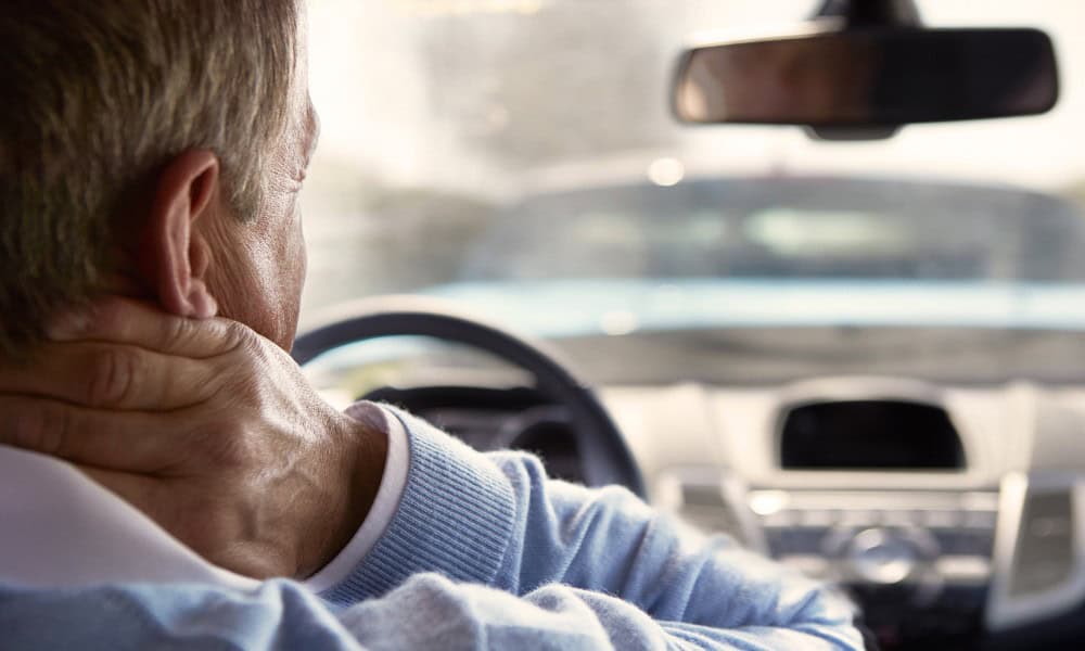 Person sitting in a car holding their neck, possibly indicating discomfort, as they look towards the windshield.