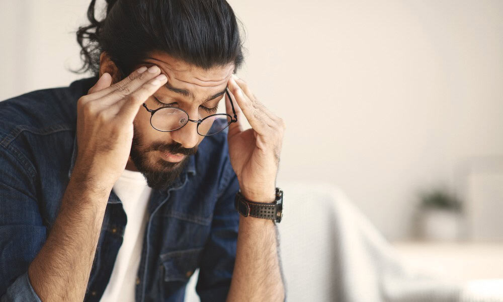 Person with glasses and a beard, wearing a denim shirt, sits and holds their temples with a pensive expression.