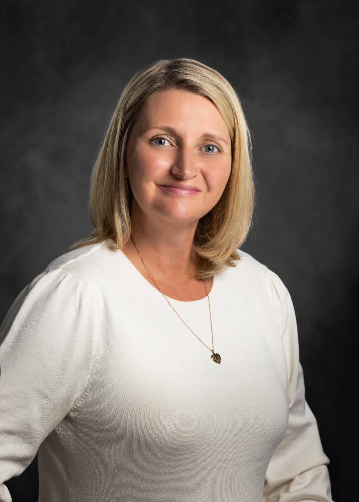 A woman with shoulder-length blonde hair is smiling and wearing a white top and necklace, against a dark background.