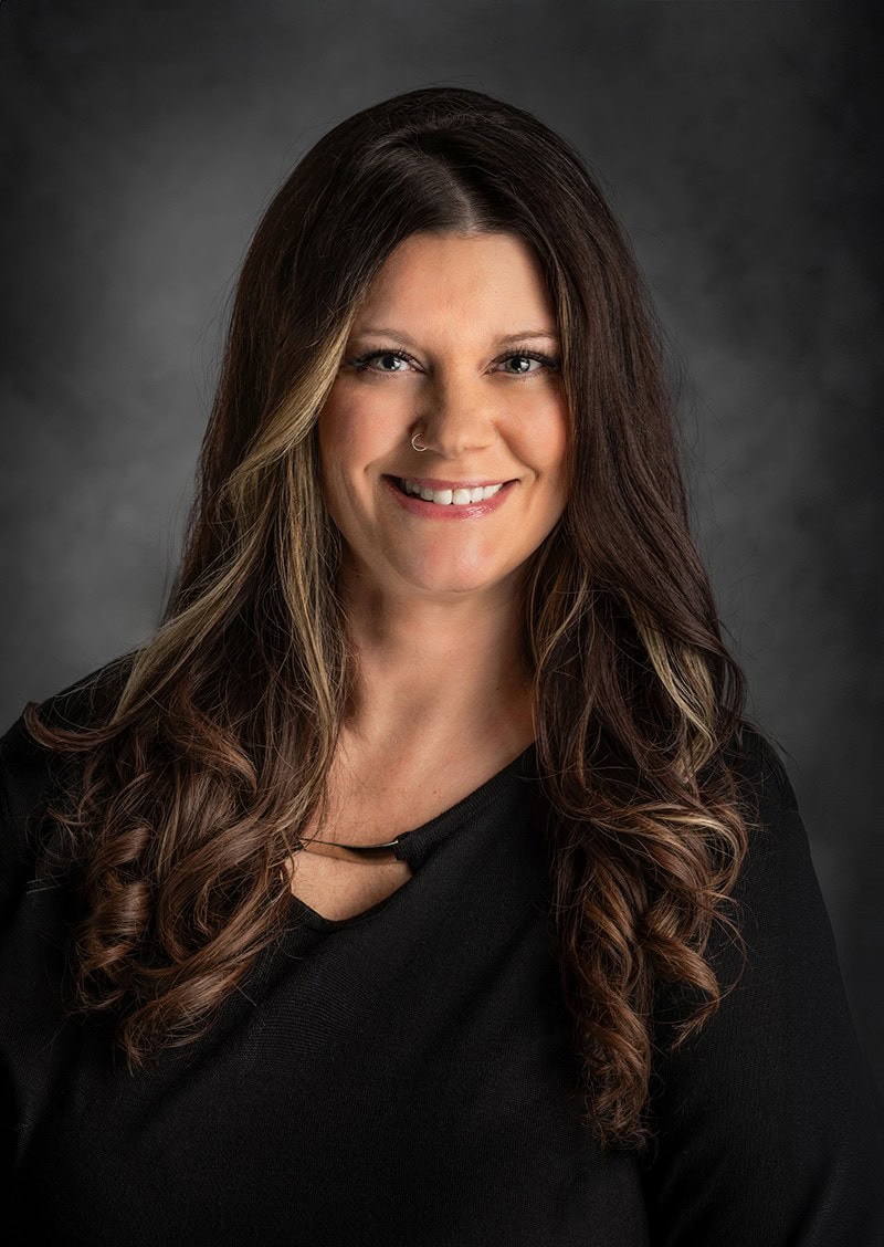 Woman with long, wavy brown hair and a nose ring, wearing a black top, smiling against a dark gray background.