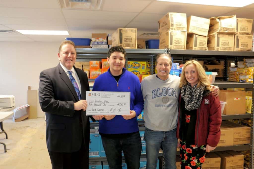 Group of four people posing in a room with shelves of boxes and snacks. One person holds a large check for $500.