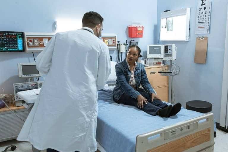 Doctor in a white coat speaks with a seated patient in a hospital room, with medical equipment surrounding them.