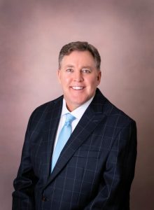 A person in a dark checkered suit and light blue tie, smiling, against a neutral studio backdrop.