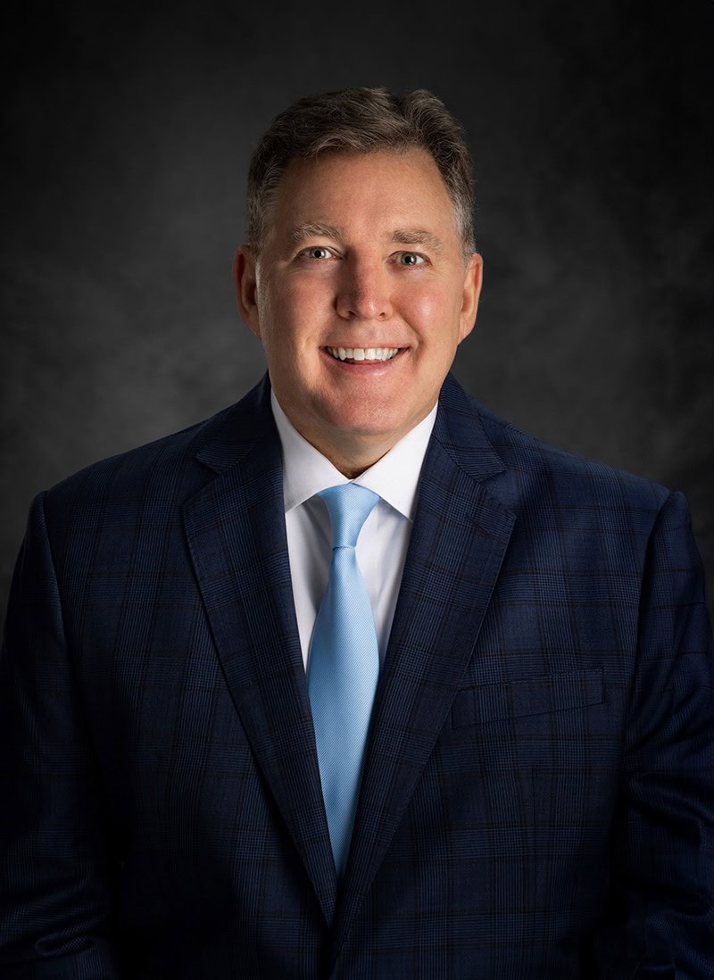 Smiling man in a dark plaid suit and light blue tie against a dark background.