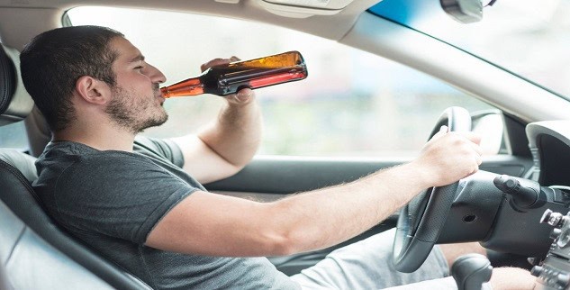 Man in a gray t-shirt drinks from a brown bottle while sitting in the driver's seat of a car.