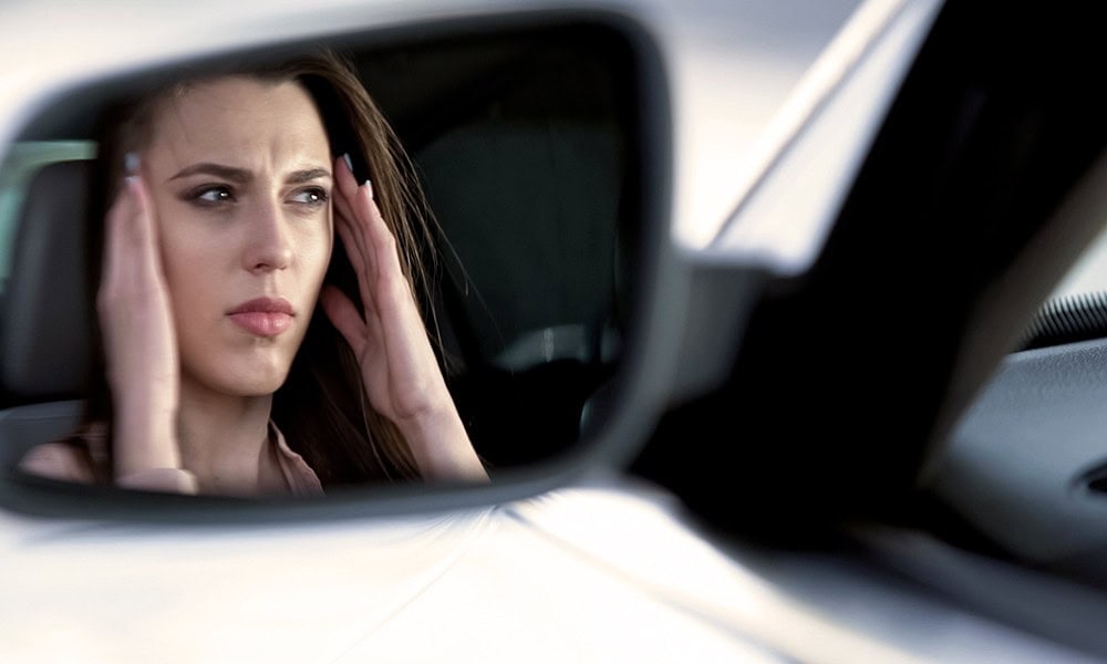 A woman is seen through a car side mirror, holding her head with both hands, looking concerned.