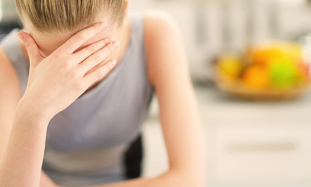 A person with blonde hair holds their head in one hand, appearing upset, with a blurred kitchen and fruit bowl in the background.