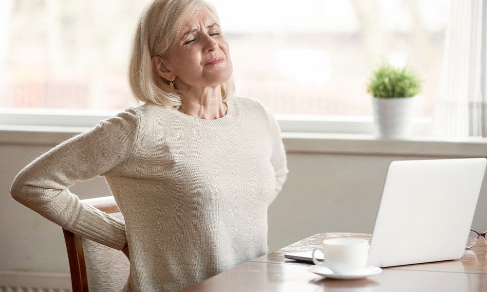 Older woman sitting at a table, clutching her lower back and grimacing. A laptop and a cup are on the table, with a potted plant in the background.