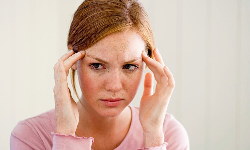 Woman with red hair touching her temples, appearing tense or thoughtful, wearing a pink shirt against a neutral background.
