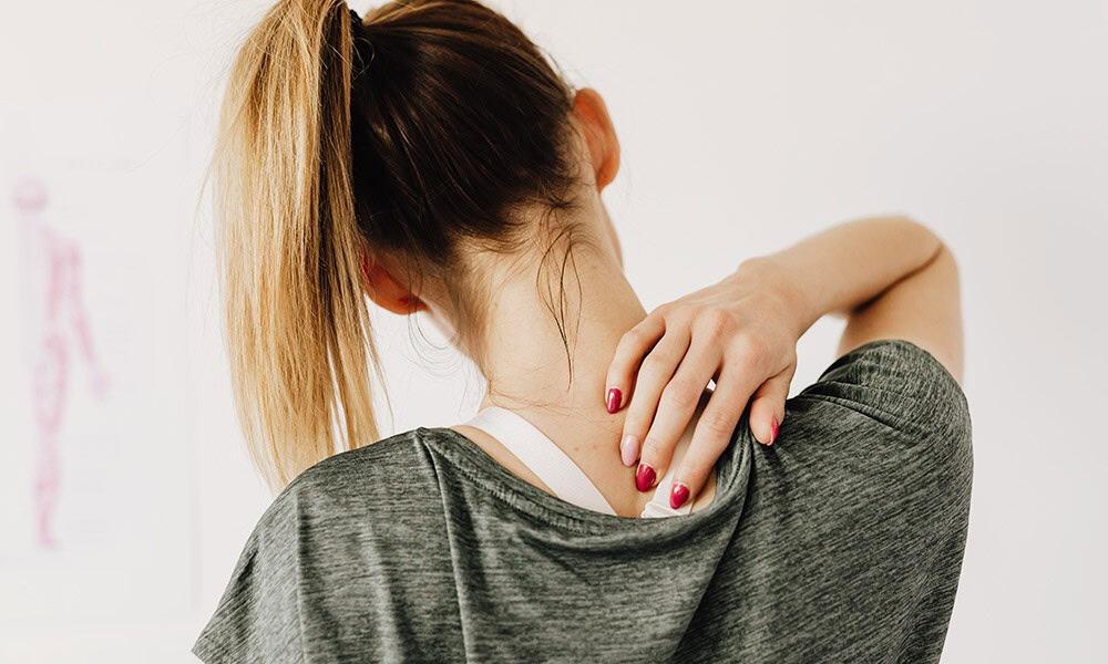 A woman with a ponytail holds her neck with her right hand, wearing a gray shirt, against a plain background.