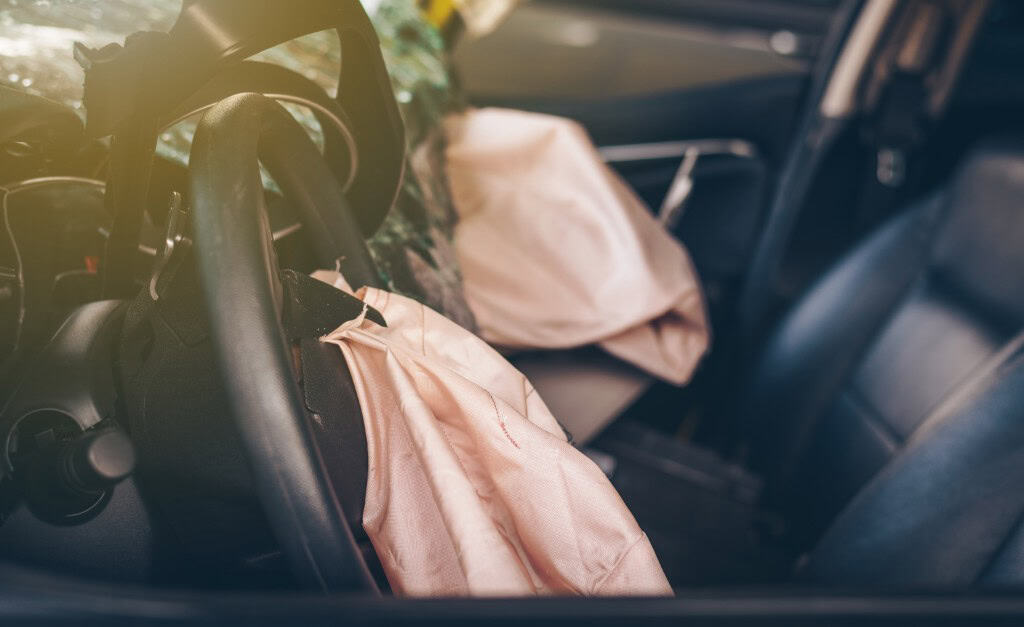 Deployed airbags in a car interior, with a sunlit steering wheel and visible tears on the fabric.