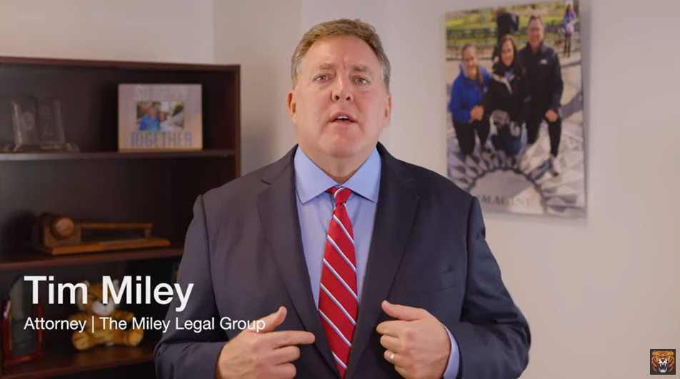 Man in a suit stands indoors speaking, with a framed family photo and books on a shelf in the background. Text overlay reads: "Tim Miley, Attorney | The Miley Legal Group.