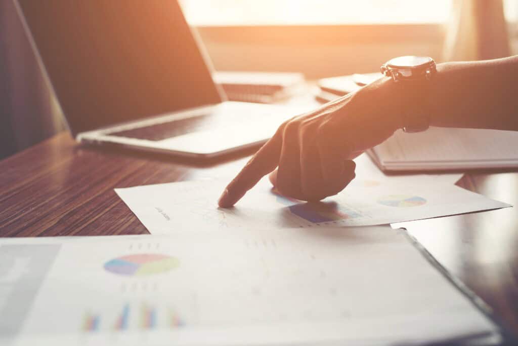 A person points at a chart on a sheet of paper on a wooden desk, with a laptop and notebook nearby.