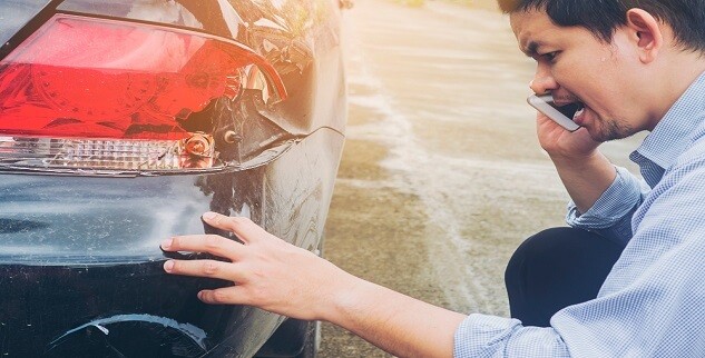 A man examines a dented car bumper while talking on a phone.