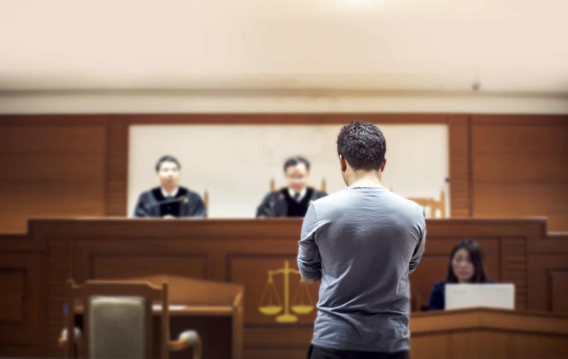 A person stands facing a courtroom, with two judges seated at the bench and a clerk writing notes.