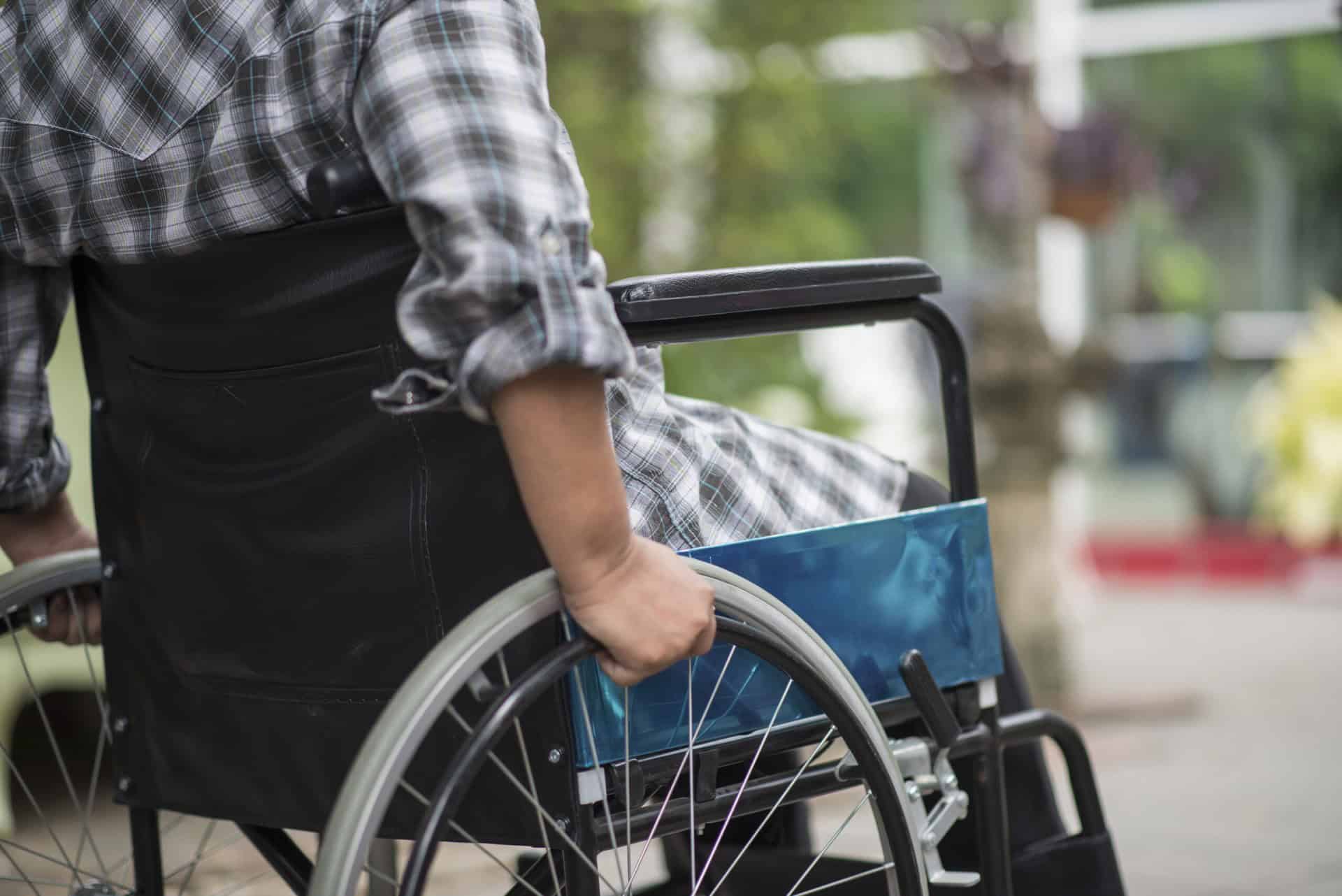 Person in a plaid shirt sitting in a wheelchair, hands on the wheels, outdoors.