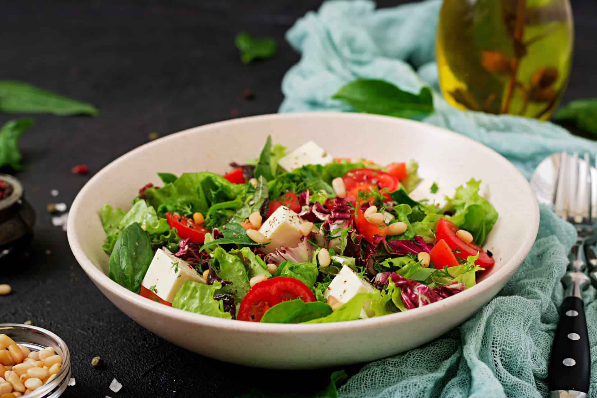 A bowl of mixed salad with lettuce, cherry tomatoes, feta cheese, pine nuts, and herbs. A bottle of olive oil and utensils are in the background.