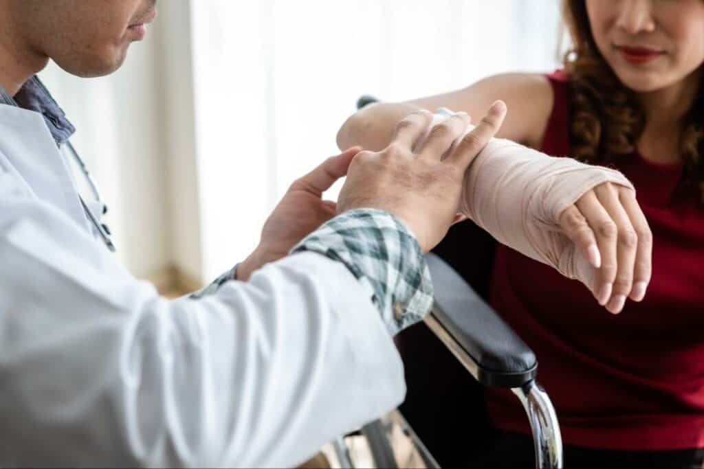 A doctor examines a woman's bandaged arm as she sits in a wheelchair wearing a red top.