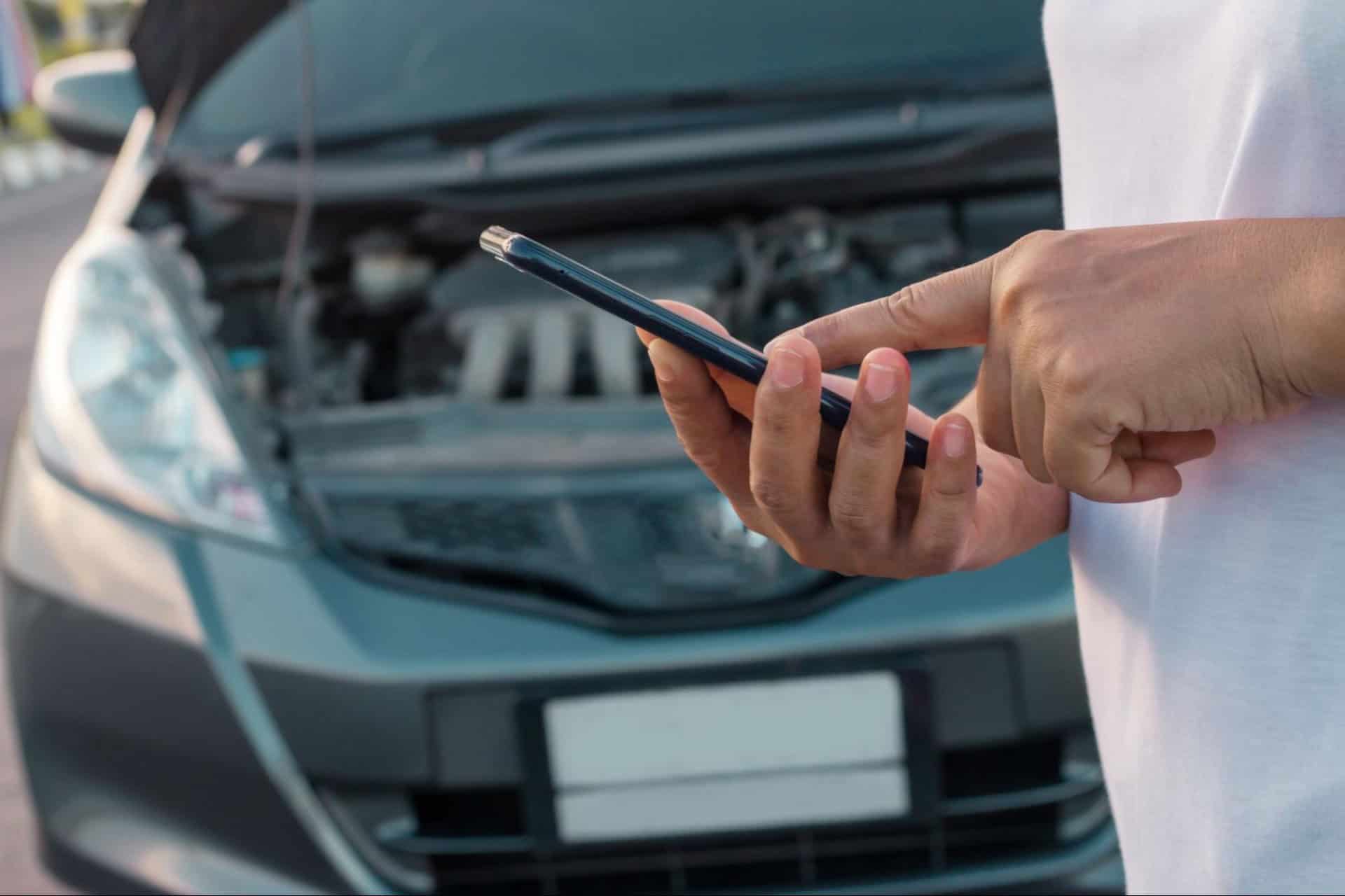 Person uses a smartphone next to a car with an open hood.