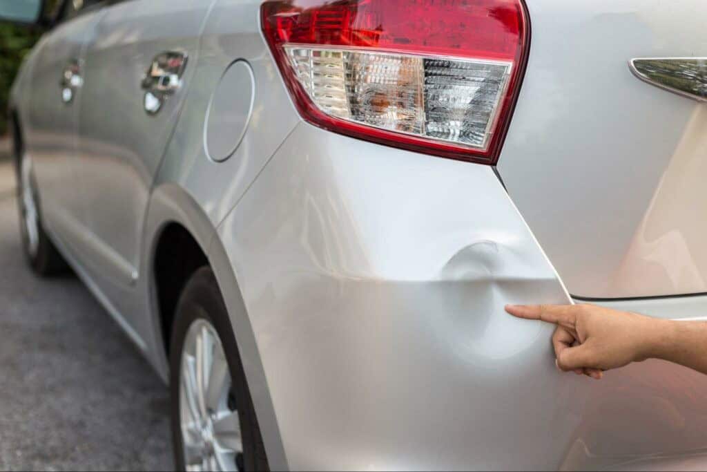 A person points to a dent on the rear right side of a silver car in an outdoor setting.