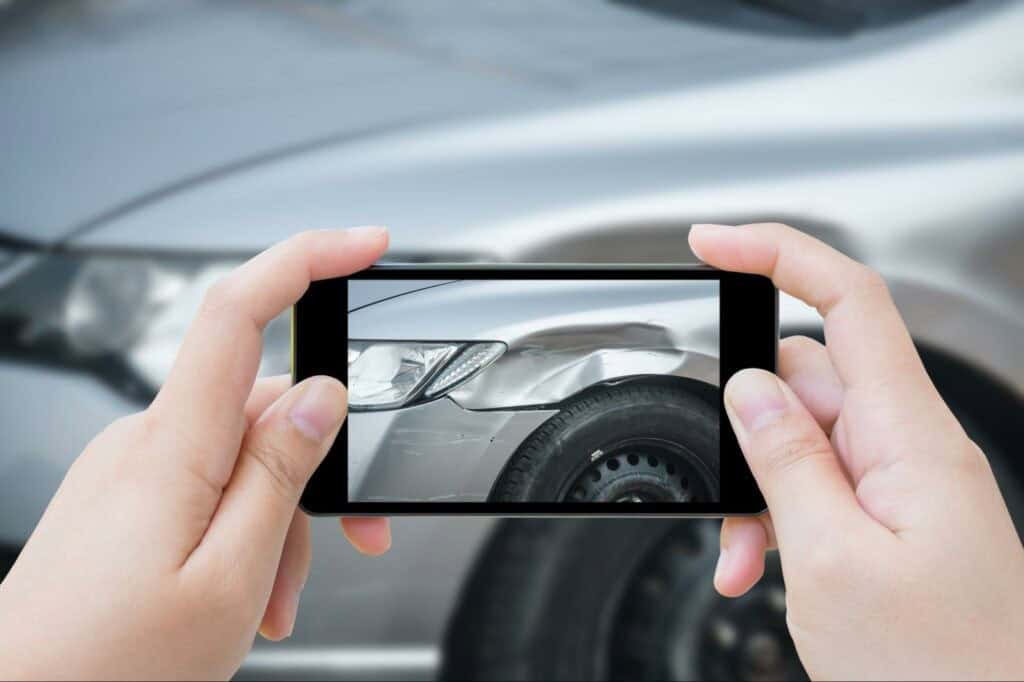 Hands holding a smartphone capturing an image of a car's dented front fender.