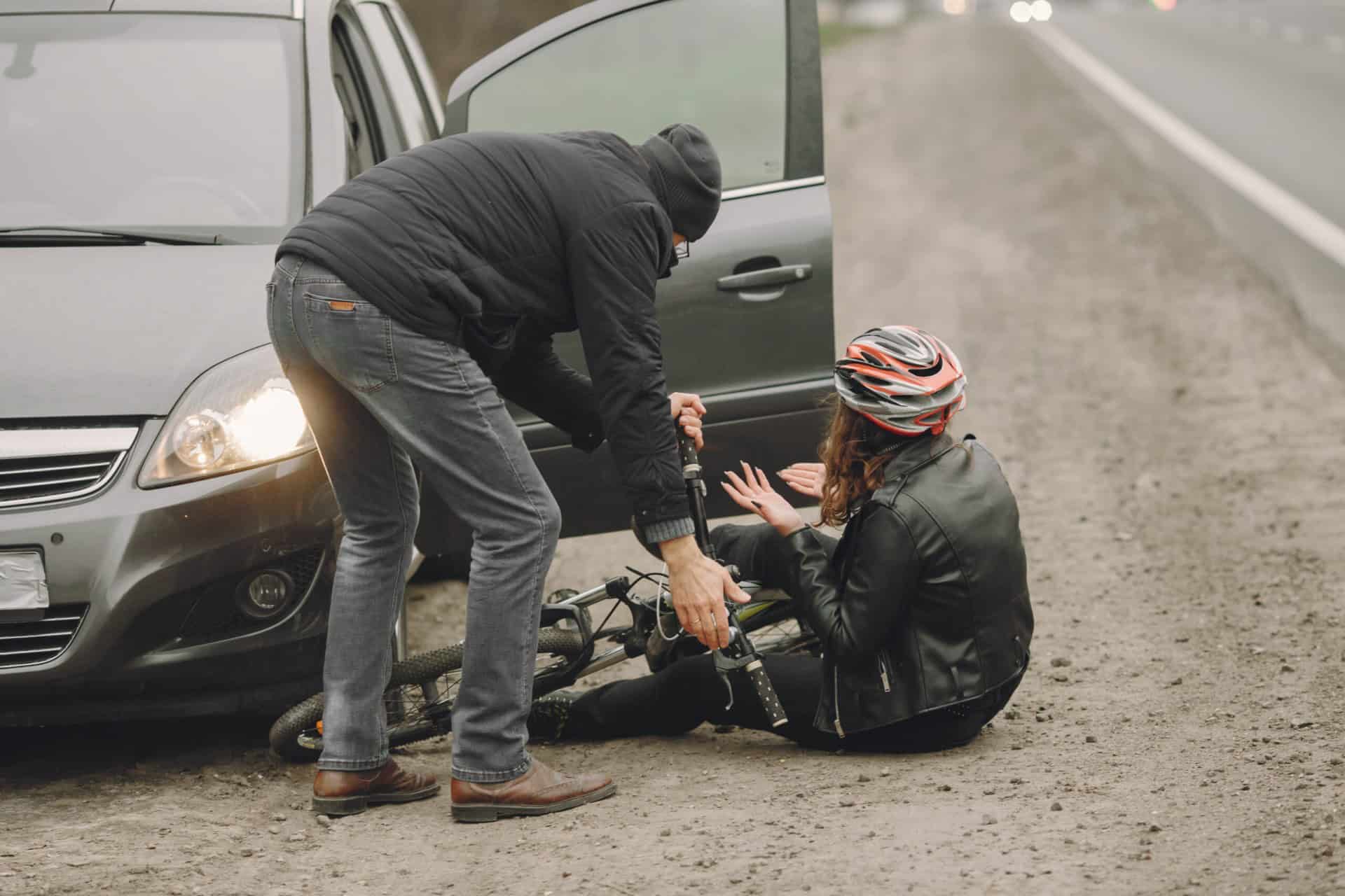 A person helps a fallen cyclist near an open car door on a roadside. The cyclist is wearing a helmet and holding the bicycle. The car is parked on a gravel path by the highway.