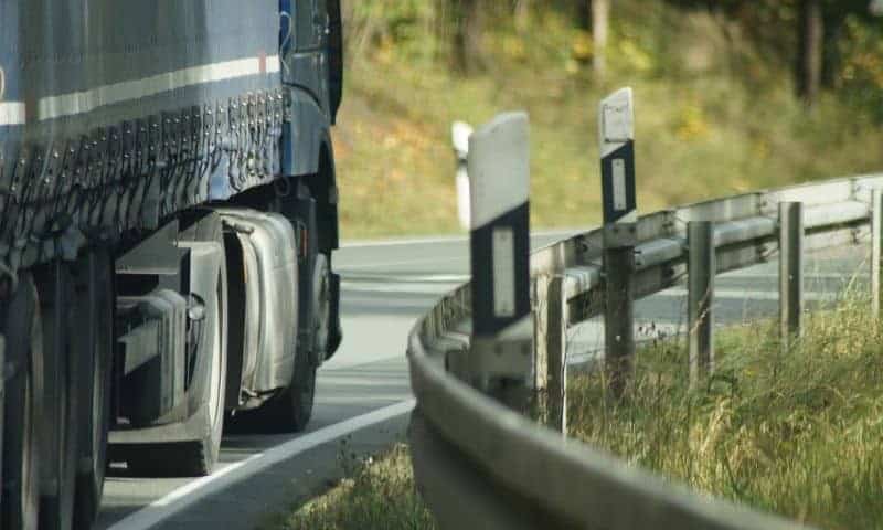 Close-up of a blue truck driving on a curved road with a guardrail and roadside markers.