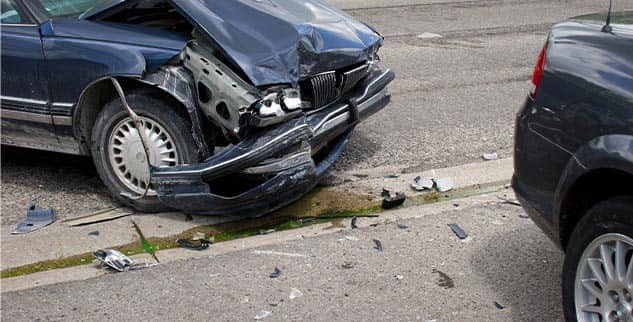 Two cars involved in a collision on a street. The front of the blue car is heavily damaged, with debris scattered on the ground. The rear of a black car is visible on the right.