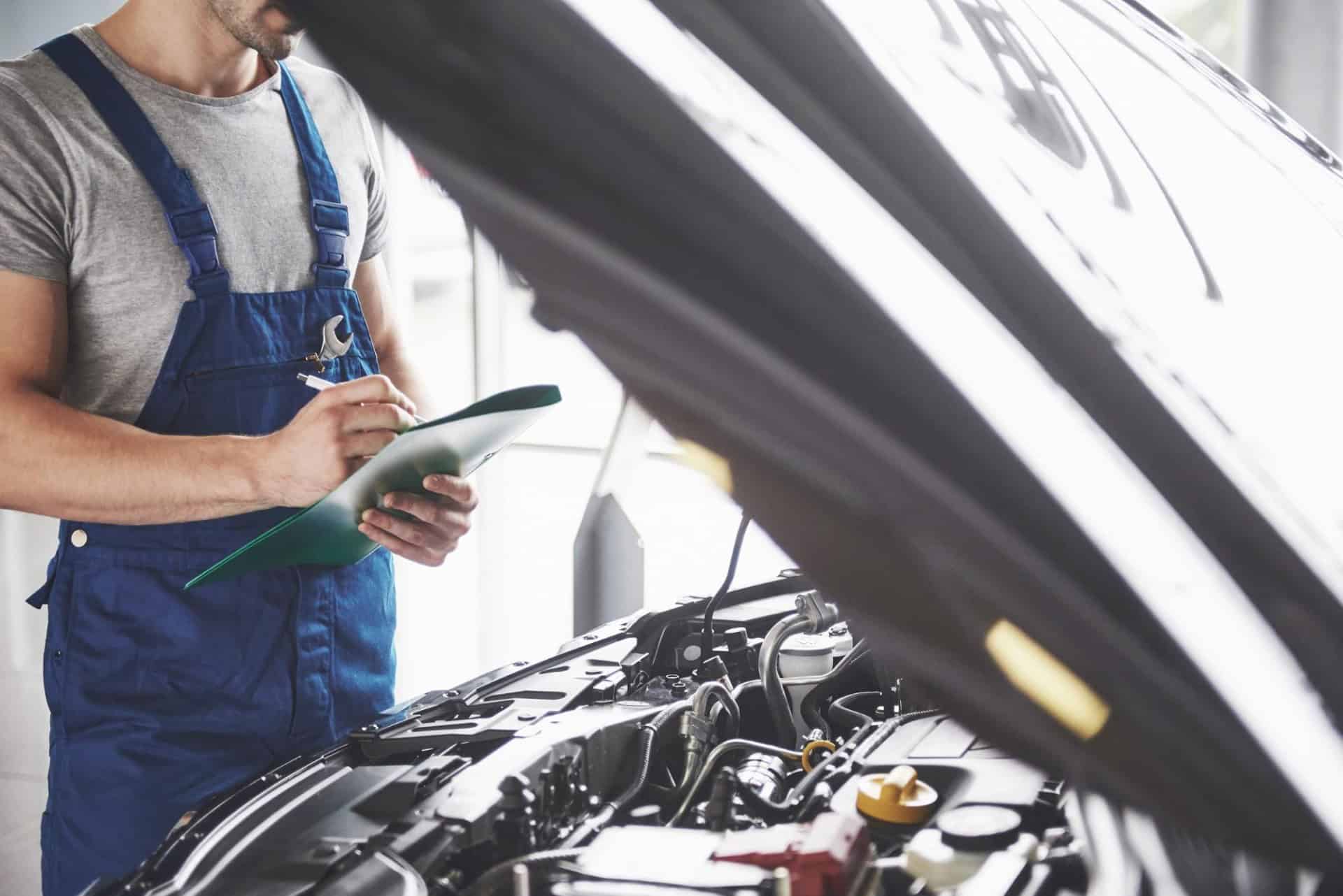 Mechanic in blue overalls examines an open car hood while holding a clipboard in a garage setting.