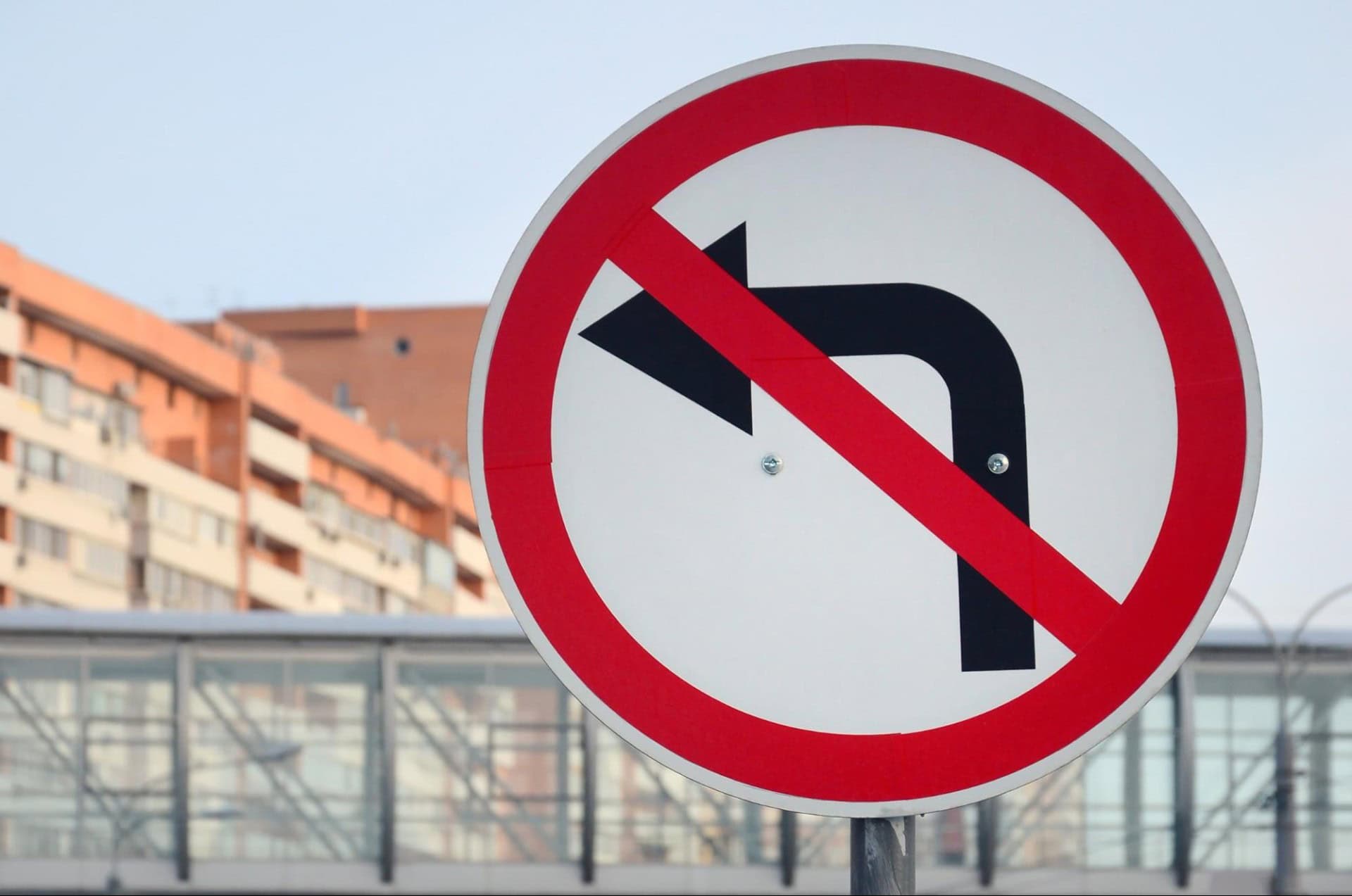 No left turn sign with a red circle and slash over a left arrow. Background shows buildings and a pedestrian bridge.