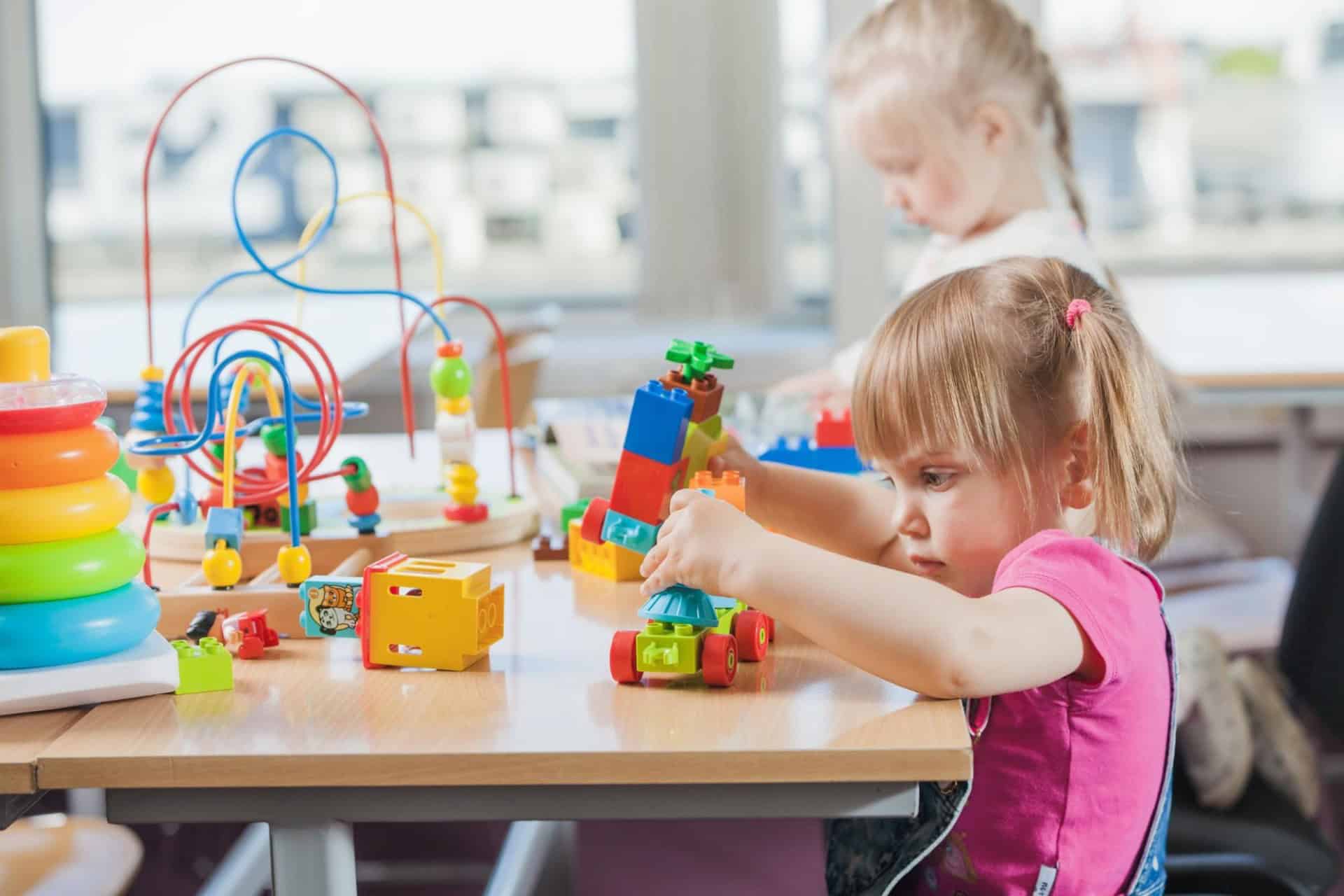 Two young children play with colorful toys and building blocks at a table in a bright room.