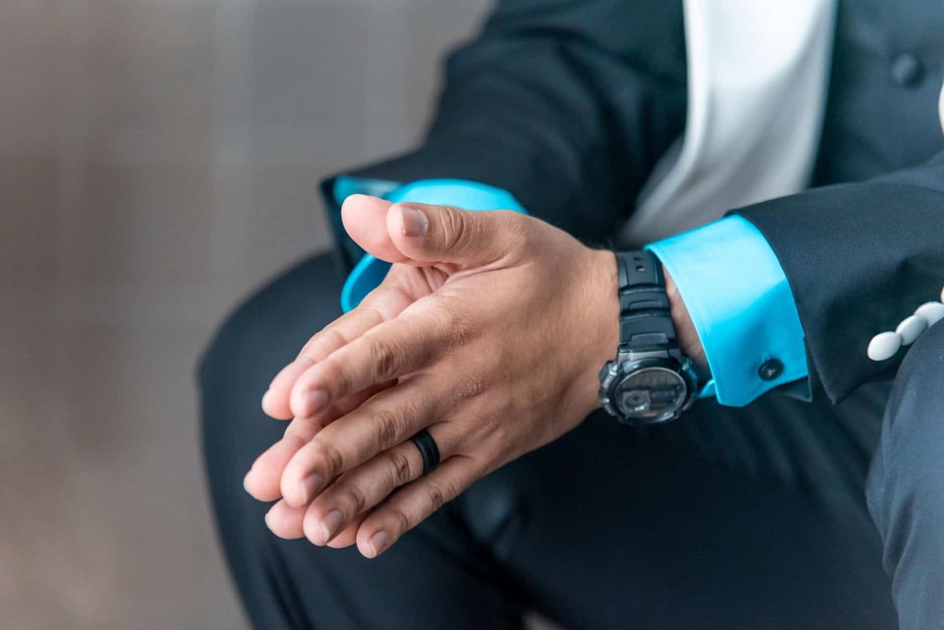 Man in a black suit with light blue shirt and a wristwatch, hands clasped together in a thoughtful pose.