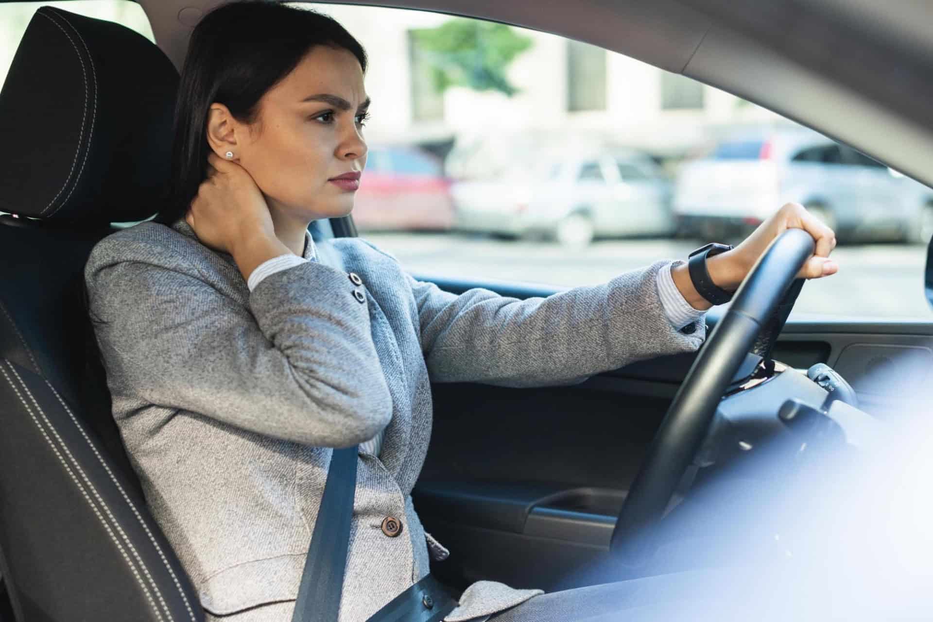 A woman in a gray blazer sits in a car, holding the steering wheel with one hand and touching her neck with the other. She looks forward with a serious expression.