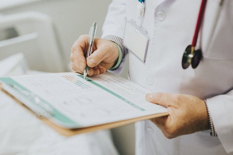 A doctor in a white coat writes on a clipboard with one hand, holding a pen. A stethoscope hangs around their neck, and a hospital bed is visible in the background.
