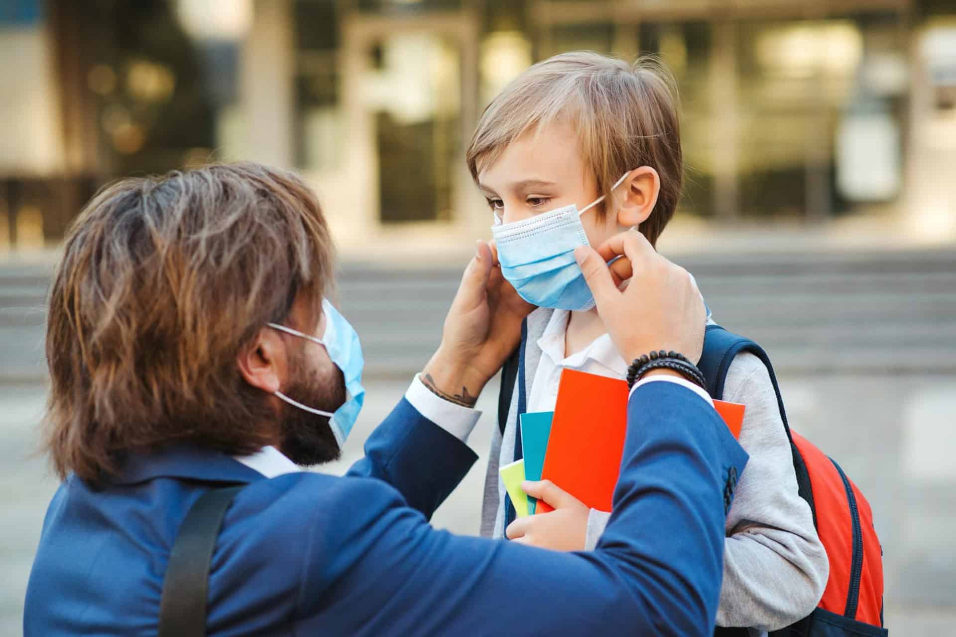 An adult helps a child adjust a face mask outdoors. The child holds books and wears a backpack. Both are wearing masks.