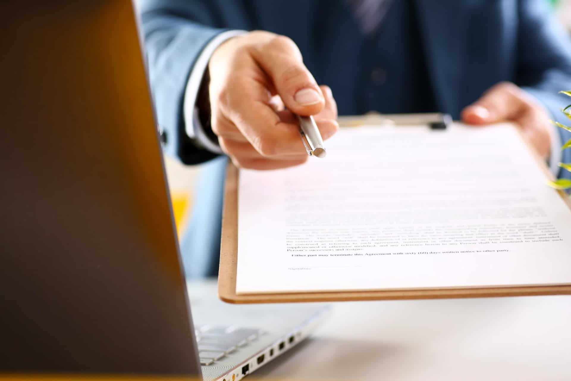 A person in a suit holds a pen and offers a clipboard with a document for signing, near a laptop.